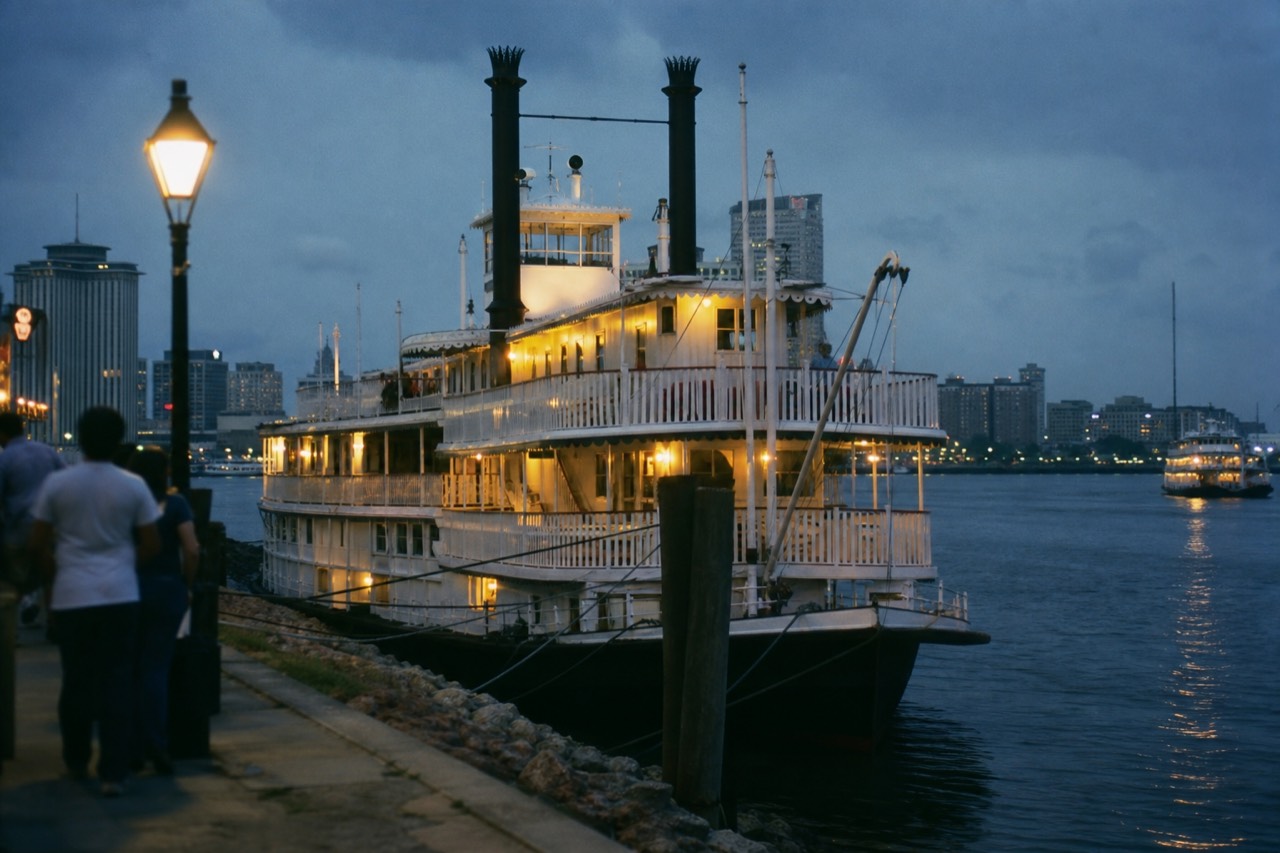 A white paddlewheel riverboat tied up at a New Orleans wharf with the Mississippi River and the curve of the downtown skyline behind it