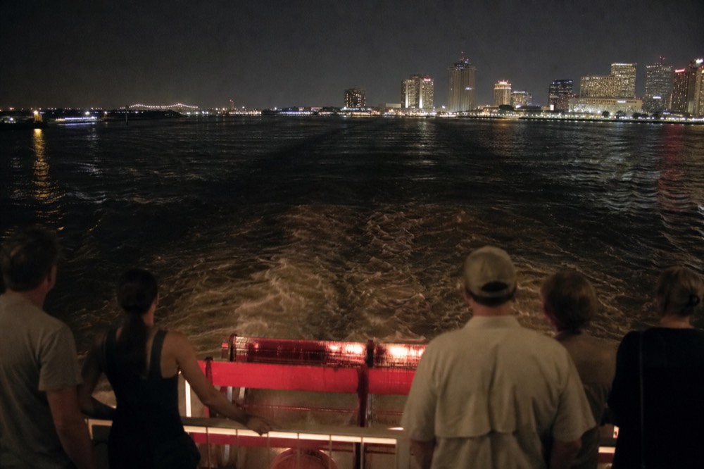 Passengers watching a red paddlewheel turn from the stern deck rail of a riverboat with the wake spreading behind