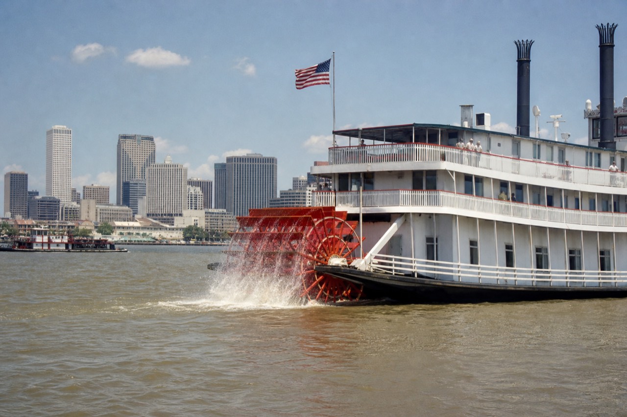 White sternwheel steamboat with twin black stacks pushing upriver past the New Orleans skyline, red paddlewheel throwing spray