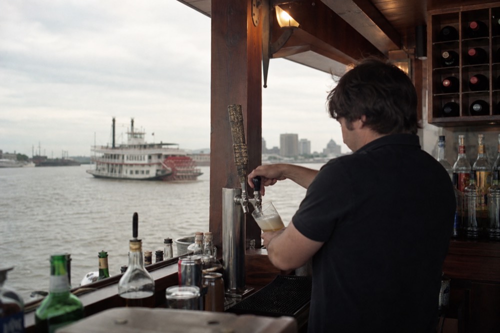Bartender pouring a beer behind a wooden riverboat bar with bottles and a wine list on the back shelf