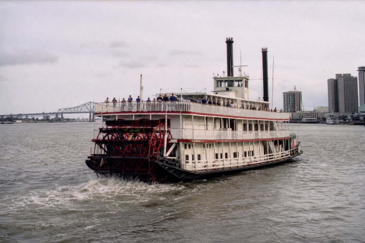 Paddlewheeler churning the Mississippi in afternoon light with passengers lining the open upper deck
