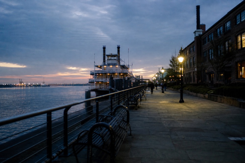 Paved riverfront walkway in New Orleans running beside the Mississippi with iron benches, a moored riverboat ahead and the brick Jax Brewery building to the right