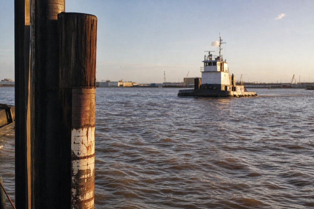 Low river gauge marker on a New Orleans wharf piling with brown water running past and a working tug beyond