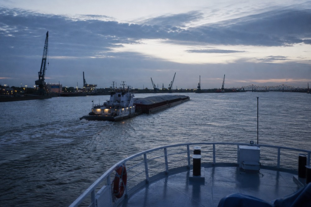 View from a riverboat bow of the Mississippi curving past container cranes and a barge tow below New Orleans