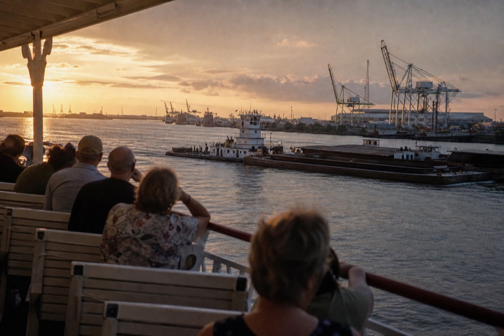 Passengers on the open upper deck of a paddlewheeler watching container cranes and barge tows along the Mississippi