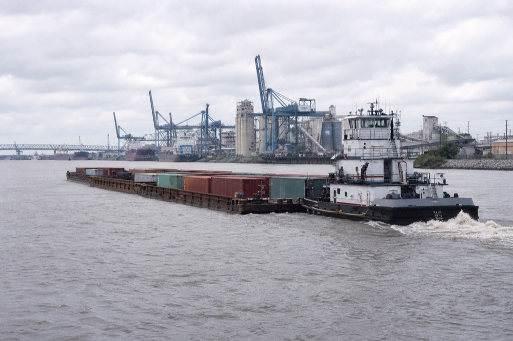 Barge tow pushed by a tug on the Mississippi with container cranes and grain elevators lining the far bank