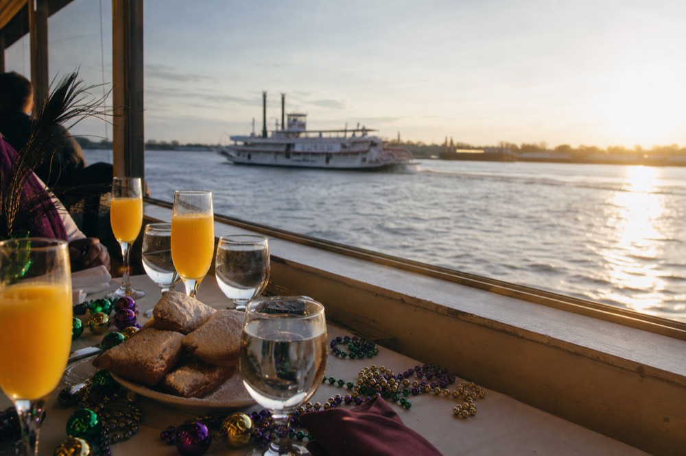 Festive brunch table on a riverboat with mimosa glasses, beignets and Carnival-coloured beads beside a window onto the Mississippi