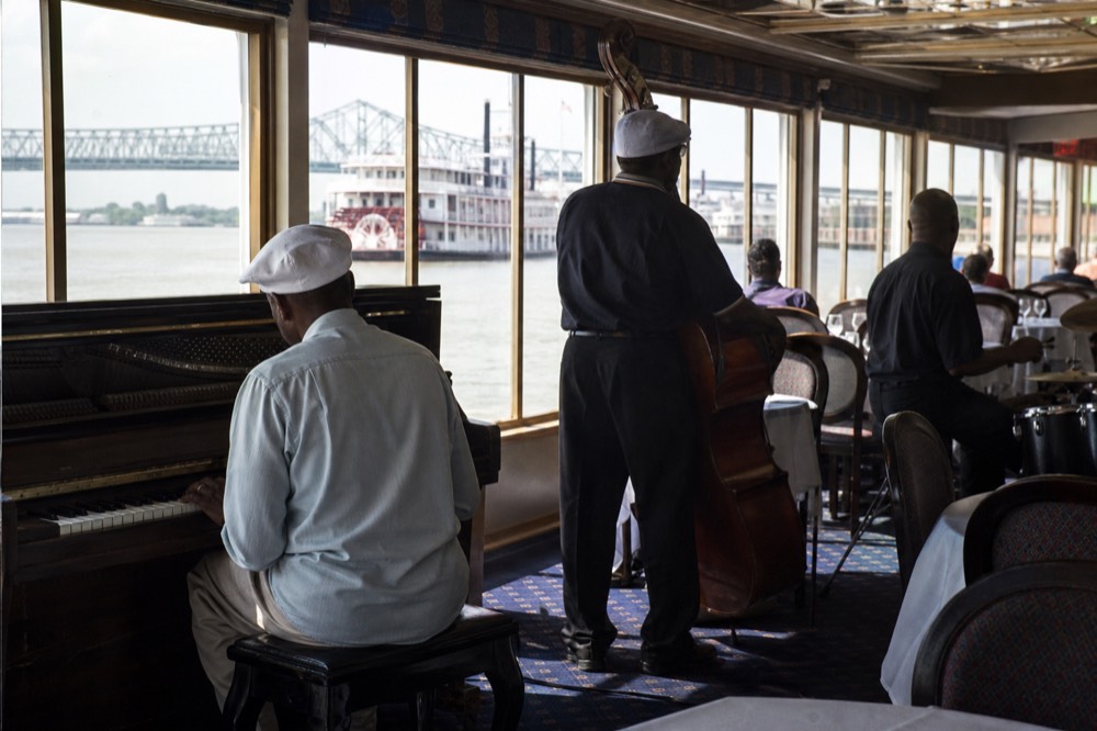 Jazz trio set up in a riverboat dining room with brass instruments and an upright piano beside tall windows