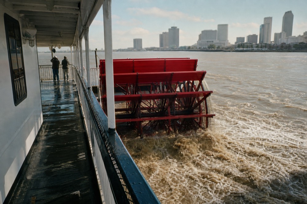 Red sternwheel paddlewheel churning brown Mississippi water behind a white excursion riverboat