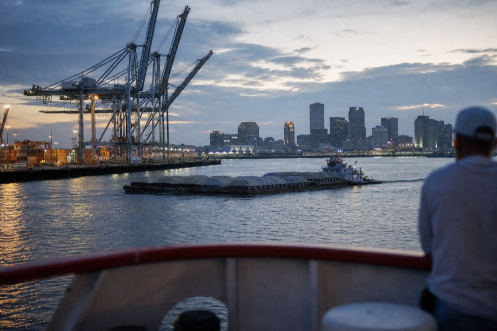 View from a riverboat deck of container cranes and a barge tow on the Mississippi with the New Orleans skyline in the background
