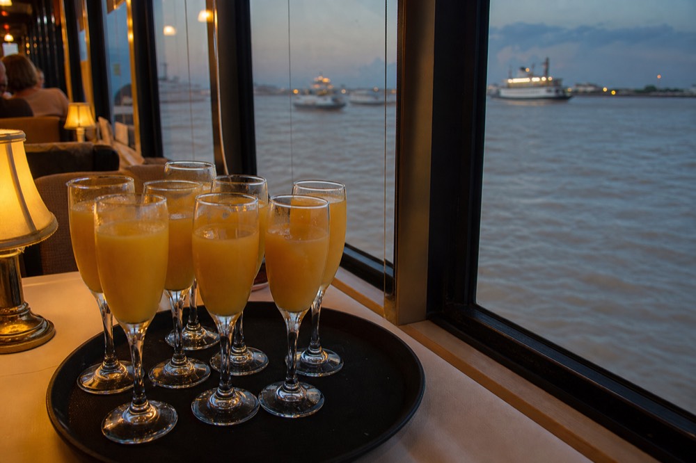 Tray of mimosas in flutes on a riverboat brunch table beside a window looking out at the wide brown river