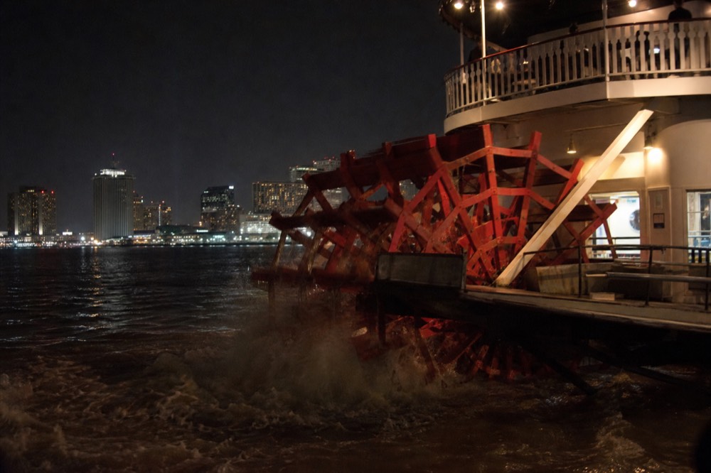 Close view of a red sternwheel churning brown river water behind a white excursion steamboat