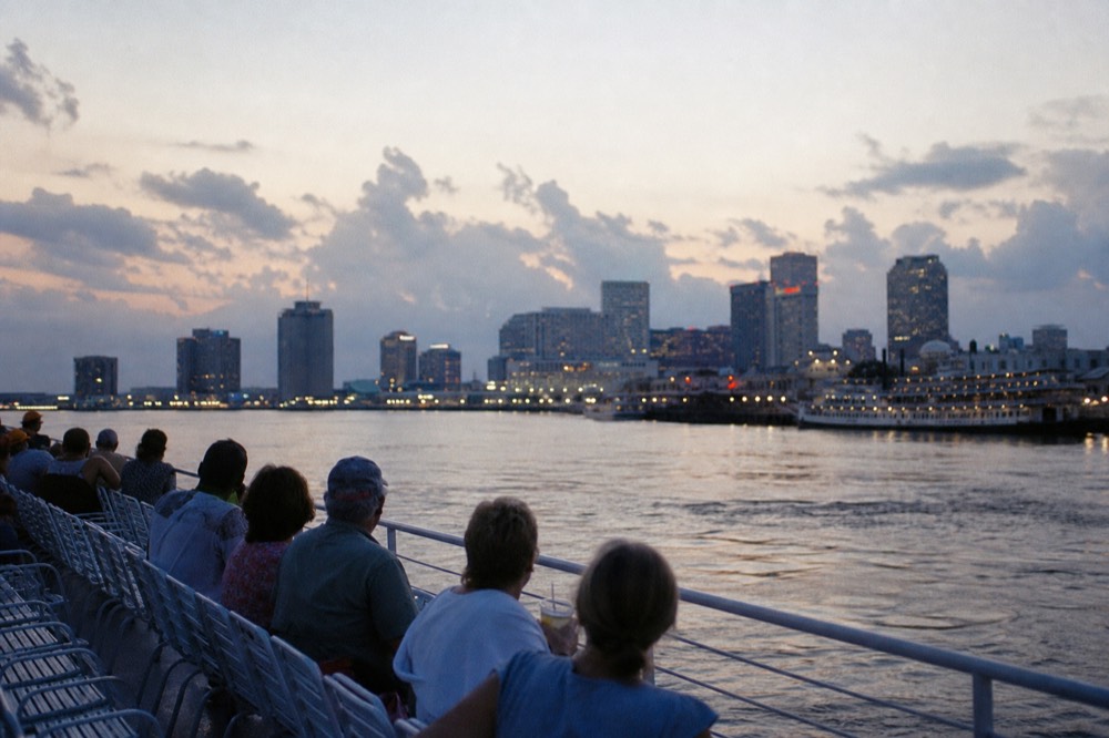 Passengers seated along the open upper deck of a riverboat at dusk with the New Orleans skyline glowing behind them