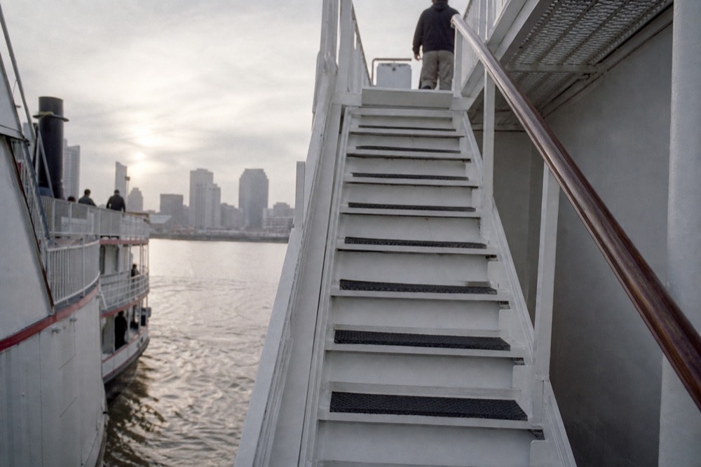 Steep white-painted steel staircase between two riverboat decks with a polished handrail and non-slip treads, river water visible through the gap