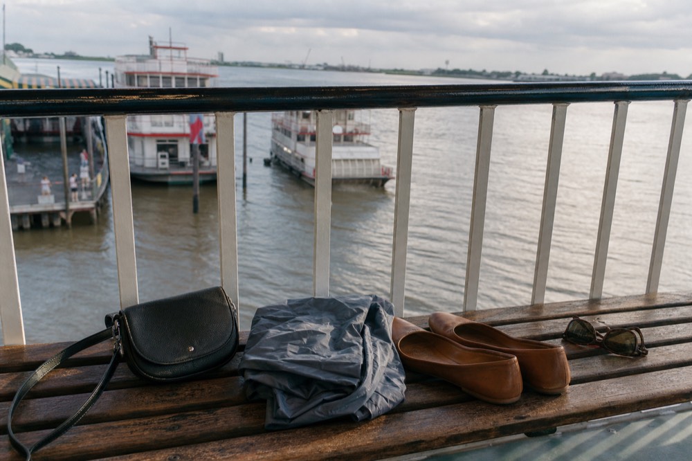 A small crossbody bag, folded rain shell, sunglasses and flat leather shoes laid out on a wooden deck bench beside a white railing